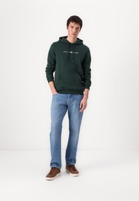 Grüne Kapuzenjacke mit weißem Text, kombiniert mit hellblauen Jeans und braunen Sneakers. Verfügt über eine Front-Kängurutasche und eine Kordelzugkapuze.