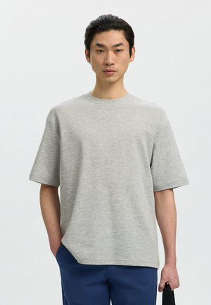 Man met zwart haar die een lichtgrijze korte mouwen shirt en blauwe broek draagt, staand tegen een effen lichte achtergrond.