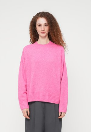 Žena s kučeravými vlasmi, oblečená v jasne ružový oversized sveter a tmavo sivé voľné nohavice, stojaca pred jednoduchým bielym pozadím.