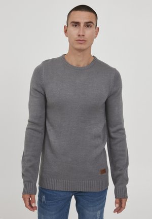 Suéter gris de punto con cuello redondo, puños y dobladillo acanalados. Presenta un tejido texturizado y un pequeño parche de piel con marca en la parte inferior.