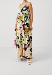 Maxikleid aus leichtem Stoff mit einem mehrfarbigen tropischen Blättermuster. Verfügt über dünne Träger und ein gestuftes Design. Getragen mit weißen Sandalen.
