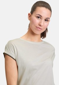 Lys beige t-shirt med rund hals og oprullede ærmer prydet med sort og guld kanting. Blød, glat tekstur.