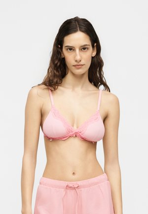 Žena s dlhými tmavými vlasmi, ktorá má na sebe ružovú podprsenku s čipkou a zodpovedajúce ružové nohavice s šnúrkou, stojaca pred jednofarebným svetlým pozadím.