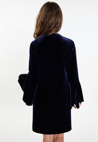 Tengeréskék bársonydressz, rézsutos hosszú ujjal és kerek dekoltázzsal, combközép hosszal és sima textúrájú anyaggal.