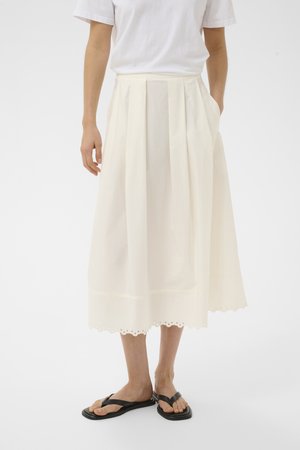 Personne portant une jupe blanche plissée mi-genou avec un ourlet festonné, une chemise blanche à manches courtes et des sandales plates noires à brides en forme de tongs.