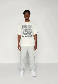 Belo-bež majica s Super Bowl grafiko, kombinirana s svetlo sivimi hlačami jogger. Obe oblačili sta iz mehkega blaga in imata sproščen kroj.