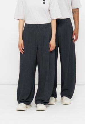 Deux personnes portant un pantalon gris foncé à jambes larges, des baskets blanches et des t-shirts blancs, debout côte à côte devant un fond blanc.
