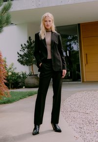 Blonde Frau in schwarzem Anzug und taupefarbenem Rollkragenpullover, die auf einem Gehweg vor einem modernen Haus mit Pflanzen und Holztür steht.