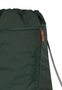 Drawstring bag in dark green fabric with a zippered pocket and leather accent. Features stitched 'Satch' logo in matching green.
