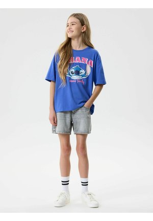 Fille aux cheveux longs portant un t-shirt bleu avec un motif Stitch, un short en jean gris, des baskets blanches et des chaussettes blanches à rayures noires, debout et souriante.