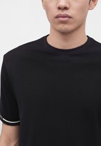 Gros plan d'une personne portant un shirt noir à manches courtes avec un col rond et un bord à rayures blanches sur la manche.