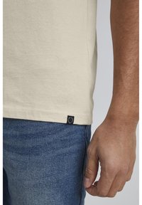 Beige bomull t-skjorte med korte ermer, som viser en liten svart etikett i kanten. Brukt sammen med blå denimjeans, som viser en teksturert stoff.