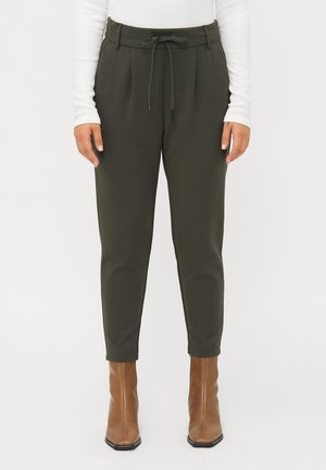 Pantaloni tapered verde oliva con pieghe frontali, vita regolabile con cordino e taglio alla caviglia. Abbinati a stivaletti marroni con tacco.