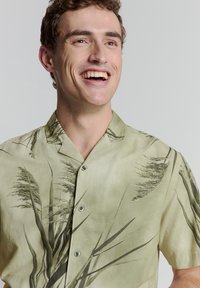 Korte mouwen, lichtgroen shirt van zacht stof, met een botanische print van donkergroene bladeren en drie knopen aan de voorkant.