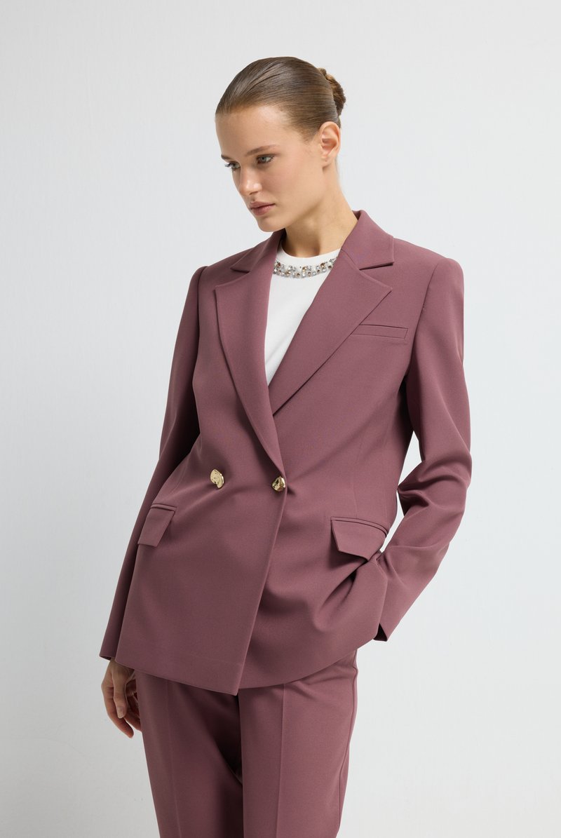 Blazer de corte cruzado en tela malva, con botones dorados, solapas pronunciadas y dos bolsillos frontales. Llevado sobre una blusa blanca con adornos.