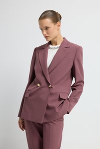 Dubbelzijdig colbert in mauve stof, met gouden knopen, scherpe revers en twee frontzakken. Draag het over een witte top met versieringen.