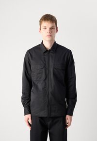 Black long-sleeve shirt with a zipper, two chest pockets, and a smooth texture. Designed with a classic collar and relaxed fit.