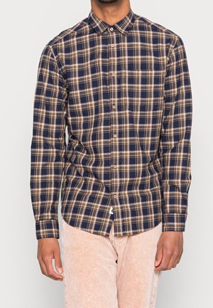 Chemise à manches longues à carreaux bleu marine et beige, avec un col boutonné, une poche sur la poitrine et un ourlet incurvé, fabriquée en tissu à carreaux.