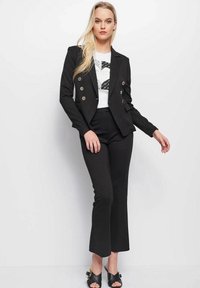Zwarte op maat gemaakte blazer met een dubbele sluiting van gouden knopen, gecombineerd met een aansluitende zwarte broek en een wit T-shirt met grafische print. Zwarte slide sandalen maken de look compleet.