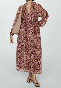 Femme portant une robe à manches longues, longue jusqu'à la cheville, avec des motifs abstraits bordeaux et beige, portant un tote bag beige et des sandales à talons noires.
