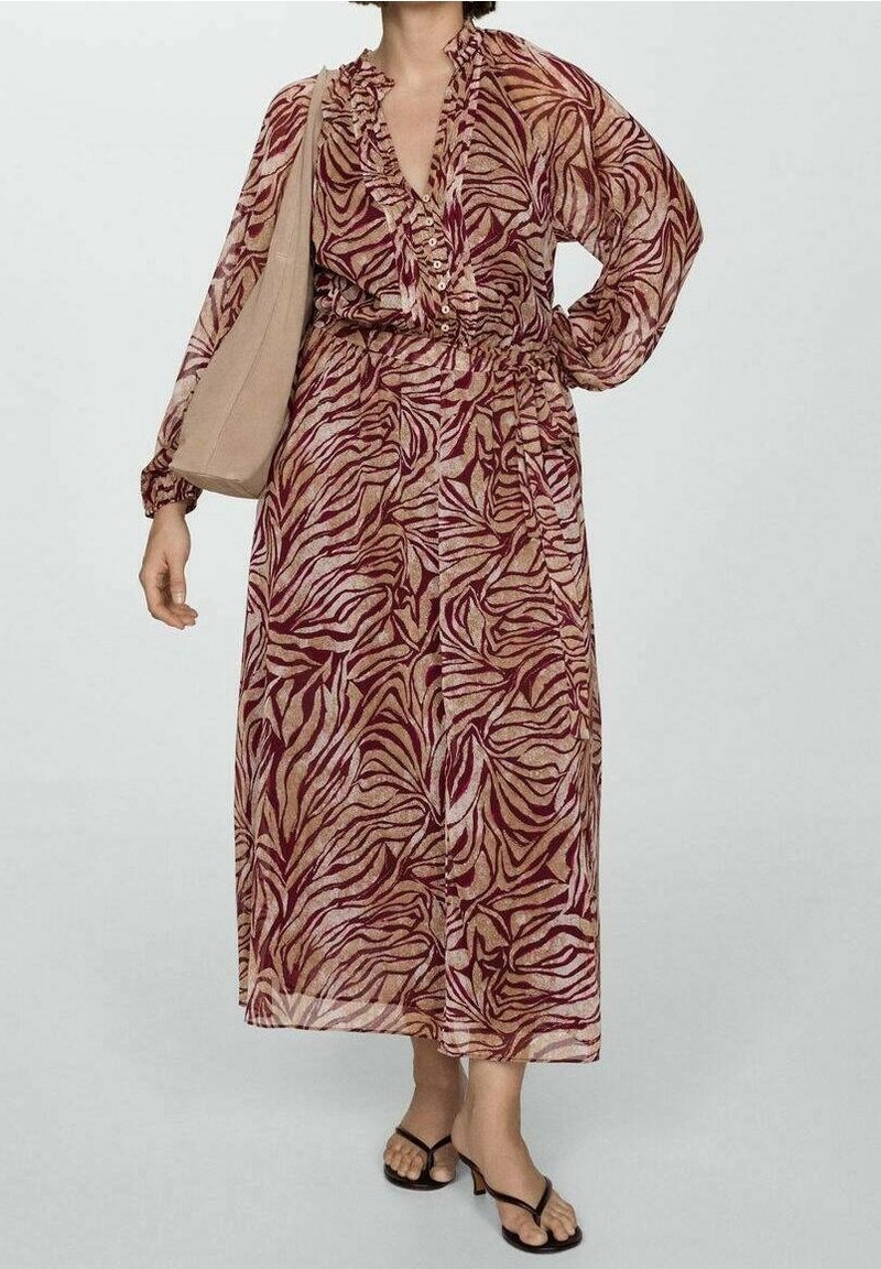 Femme portant une robe à manches longues, longue jusqu'à la cheville, avec des motifs abstraits bordeaux et beige, portant un tote bag beige et des sandales à talons noires.