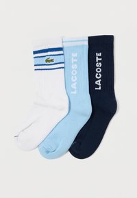 Trei perechi de șosete din bumbac: albe cu dungi albastre și logo-ul unui crocodil verde, bleu deschis și bleumarin închis, având textură ribată și logo.