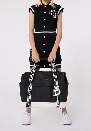 Osoba w czarnej sukience zapinanej na guziki i białych trampkach trzyma dużą czarno-białą torbę typu tote marki Karl Lagerfeld z paskami z logo i breloczkiem w kształcie kota.