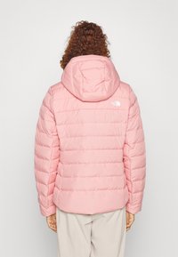 Veste rose rembourrée avec capuche, comportant des sections matelassées et un logo blanc au dos. Texture lisse et silhouette moderne.