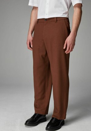 Braune gewebte Hose mit lockerer Passform, ausgestattet mit einem Reißverschluss, einem einzelnen Knopfverschluss und vorderen Taschen, kombiniert mit schwarzen Slippern.