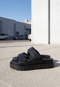 Sandales à plateforme noires avec semelles épaisses et brides réglables, de marque "BUFFALO", posées sur un trottoir en béton près d'un mur en briques blanches.