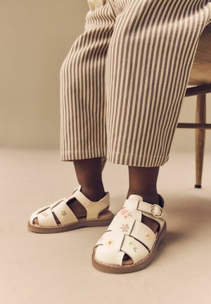 White leather sandals with floral embroidery, open-toe design, strap closure, and a brown sole. Striped beige trousers.