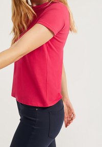 Žena s dlouhými blond vlasy má na sobě jasně růžové tričko s krátkým rukávem a tmavě modré kalhoty na jednobarevném světlém pozadí.