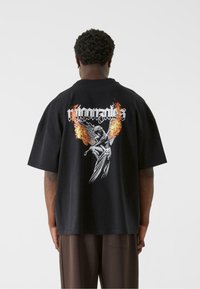 Sort bomulds T-shirt med løs pasform, der har et grafisk motiv af en engel omgivet af flammer på bagsiden og fed tekst over designet.