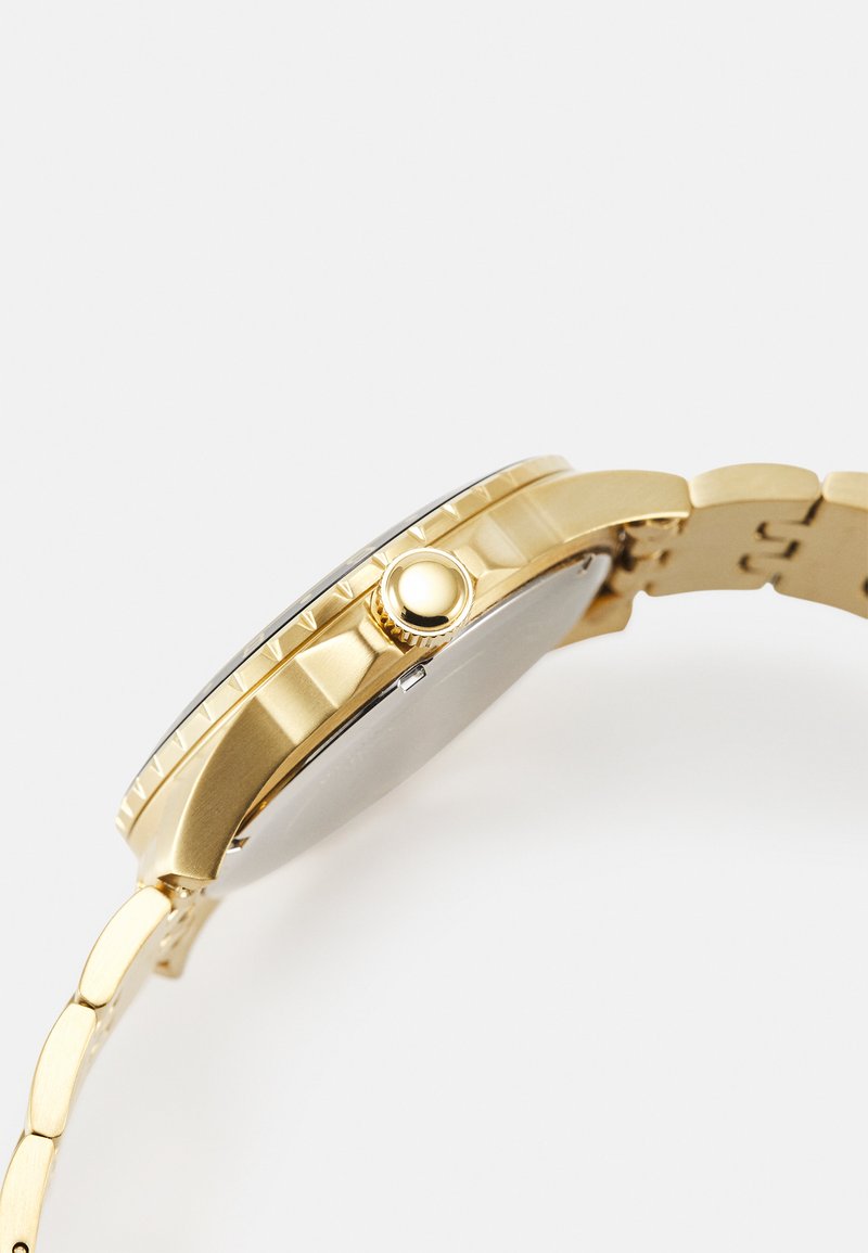 Reloj de metal dorado con esfera redonda, corona en el lateral y pulsera de eslabones. Presenta un acabado pulido y detalles de textura en el bisel.