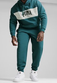 Sudadera color verde azulado con una franja horizontal blanca, que presenta el logo de "PUMA" y pantalones de chándal a juego. Zapatillas blancas completan el conjunto.