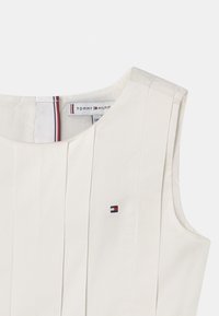 Ärmlös topp i krämfärg med vertikala veck, prydd med en liten broderad logotyp i röd, vit och blå på bröstet.