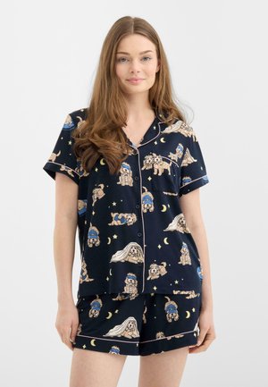 Ung kvinde iført marineblåt kortærmet pyjamassæt med knapper og mønstre af tegneseriehund og måne, stående mod en ensartet lys baggrund.