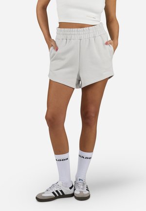 Pantalonii scurți de culoare gri deschis, dintr-un amestec de bumbac, au un elastic în talie, buzunare laterale și o croială lejeră. Sunt asortați cu șosete albe și adidași gri.