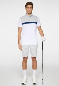 White polo shirt with gray shoulder panels and a navy horizontal stripe, paired with light gray shorts, white sneakers, holding a golf club.