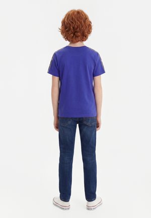 Bambino con capelli ricci rossi in piedi di spalle, indossa una maglietta blu, jeans scuri e scarpe da ginnastica bianche su sfondo bianco.