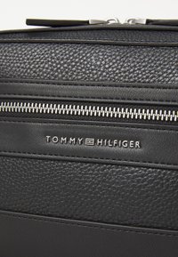 Borsa in pelle nera con superfici texture, cuciture orizzontali, una cerniera argentata e una piastra con logo metallico recante la dicitura "TOMMY HILFIGER."