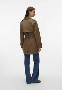 Brun trenchcoat med bälte i midjan, axelklaffar och en medellång design, tillsammans med blå utsvängda jeans och svarta ballerinas.