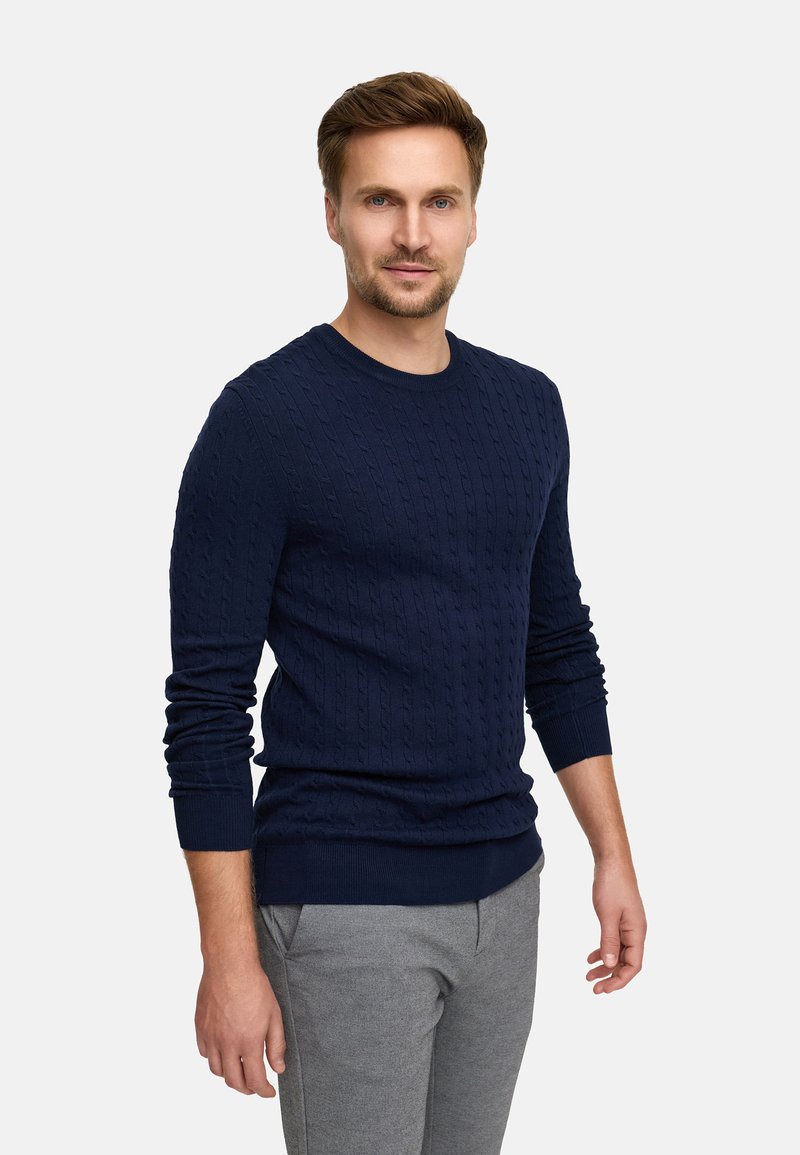 Granatowy sweter w splot kablowy z długimi rękawami i okrągłym dekoltem, w zestawie z szarymi spodniami, posiadającymi ściągacz u dołu i na mankietach.