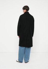 Cappotto lungo nero con taglio dritto, dotato di colletto classico e design minimalista, abbinato a pantaloni in denim blu svasati.