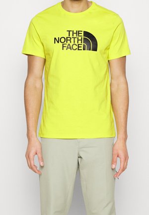 Personne portant un T-shirt jaune vif à manches courtes avec le logo « THE NORTH FACE » en noir, associé à un pantalon beige clair.