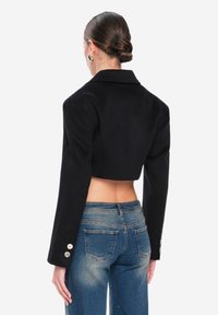 Blazer negro recortado con cuello y mangas largas, con botones dorados como detalle y combinado con jeans vaqueros azules desgastados.