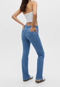 Jean en denim bleu clair flare à taille haute, avec cinq poches, et une texture qui semble douce. Porté avec un haut blanc ajusté, vu de dos.