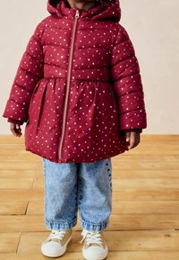 Abrigo acolchado rojo con patrón de estrellas doradas, cremallera frontal y capucha. Llevado sobre pantalones de mezclilla azules y zapatillas con acentos dorados. Fondo de suelo de madera.