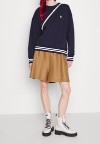 Granatowa bluza z białymi paskami, szerokie beżowe szorty oraz białe botki z czarnymi podeszwami. Na piersi znajduje się małe, haftowane logo.