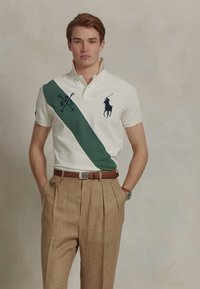 Mladenič v beli polo majici z zeleno diagonalno črto in rjavimi karirastimi hlačami, ki stoji s hands in žepih na nevtralni podlagi.
